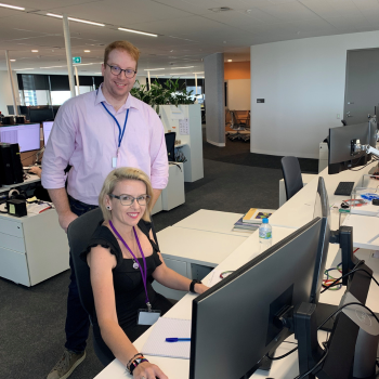 Two colleagues in a modern office, smiling at the camera. One is seated at a desk with multiple monitors, while the other stands behind.